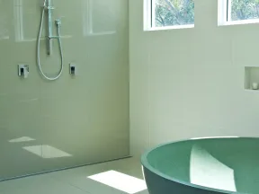 Bright modern bathroom with a green freestanding tub, wall-mounted shower, large windows, and beige tile walls.