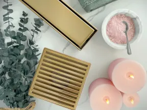 Gold shower drain grate with pink candles, eucalyptus branches, green towel, and bowl of pink bath salts on marble.