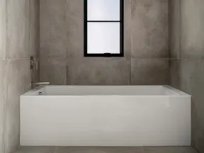 Minimalist bathroom with a modern rectangular white bathtub, grey tiled walls, and a tall frosted window.