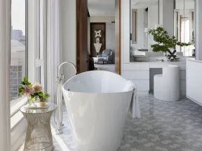 Modern bathroom with freestanding white tub, floral side table, large mirror, and city view through windows