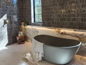 Modern bathroom with dark gray tiled walls, freestanding gray tub, marble accents, and leaf-shaped chandelier.