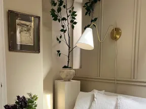 Cozy corner with white couch, three pillows, wall lamp, potted plant, dark wooden table, and framed artwork on beige walls.
