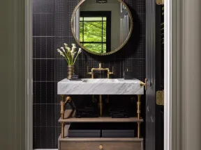 Modern bathroom with marble sink, gold fixtures, round mirror, and black tiled wall with decorative patterns.