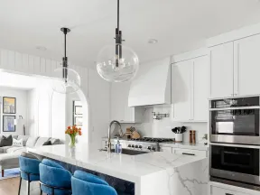 Modern white kitchen with marble island, blue velvet bar stools, pendant lights, and stainless steel appliances