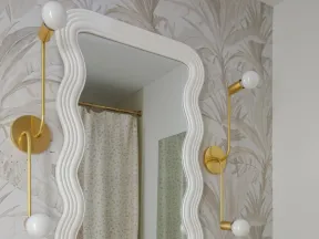 Elegant bathroom with wavy white mirror, gold fixtures, floral wallpaper, and beige marble countertop with sink.