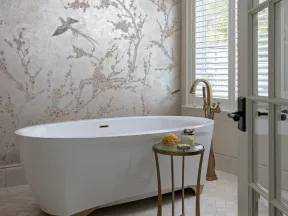 Elegant bathroom featuring a white freestanding tub, gold fixtures, floral wallpaper, and arched window with shutters.