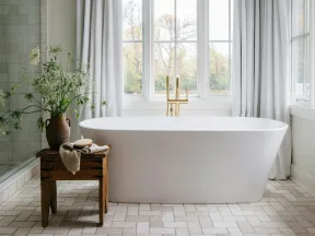 Bright bathroom with freestanding white bathtub, wooden stool, vase of flowers, and large window with white curtains.