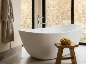 Modern white freestanding bathtub next to large windows with natural scenic view and wooden stool with bath sponge.