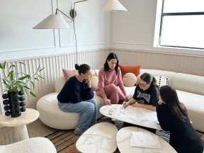 Four women collaborating over architectural plans in a modern, bright living room with contemporary furniture.