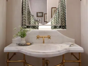 Elegant bathroom sink with gold fixtures, oval mirror, patterned curtains, wall sconces, and decorative vase.