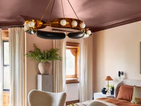 Cozy bedroom with beige armchairs, rust-colored bedding, large windows, and modern chandelier lighting.