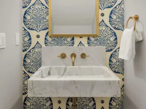 Marble sink with gold fixtures, ornate gold-framed mirror, blue and white patterned wallpaper, and gold wall sconces.