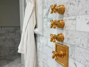 White bathrobe hanging next to four gold vintage shower controls on a marble tiled wall in a bathroom.