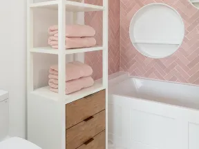 Modern bathroom with white shelving unit holding pink towels, wood drawers, pink tiled wall, and white bathtub.