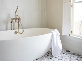 Modern white freestanding bathtub with chrome faucet and white towel beside window on patterned tile floor.