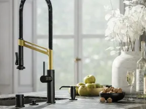 Modern kitchen faucet with black and brass finish on marble countertop near bowl of walnuts and green apples.