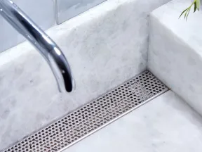 Close-up of modern chrome faucet and wall-mounted handle over white marble sink with metal drain cover.