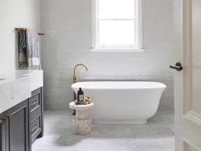 Modern bathroom with freestanding white tub, brass faucet, gray cabinets, marble floor, and a window above bathtub.