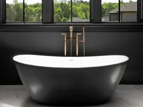 Modern black freestanding bathtub with gold faucet under windows showing green outdoor trees.