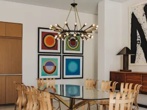 Modern dining room with glass table, unique wooden chairs, abstract wall art, and a contemporary chandelier.