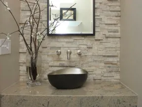 Modern bathroom sink area with stone wall, black vessel sink, rectangular mirror, and decorative vase with branches.