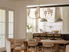 Modern dining room with wooden table, beige chairs, gold chandelier, and open kitchen with bar stools.