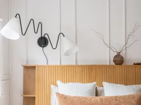Cozy modern bedroom with wooden headboard, white bedding, textured pillows, wall-mounted zigzag lamp, and decorative vase.
