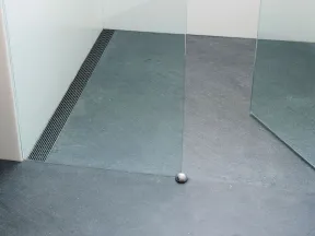 Modern minimalistic shower area with gray stone floor, glass partitions, and a metal drain along the corner.