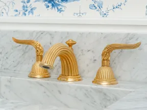 Elegant gold faucet with winged handles on a marble sink under a gold-framed mirror and blue floral wallpaper.