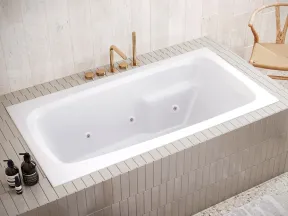 Modern white built-in bathtub with golden fixtures in a beige tiled bathroom with wicker basket and wooden chair.