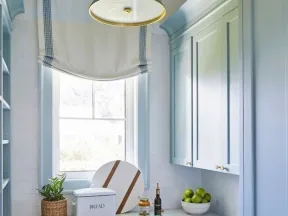 Bright kitchen with blue cabinets, white countertops, window with Roman shade, and countertop decor including bread box and fruit bowl.