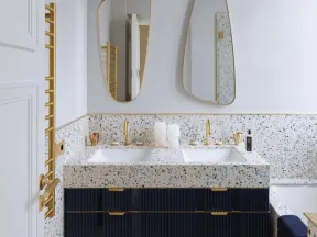 Modern bathroom with terrazzo double sink, gold fixtures, navy cabinets, two asymmetrical mirrors, and white wall lights.