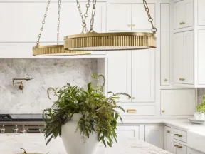 White kitchen with marble countertops, brass pendant lights, green plant centerpiece, and fruit bowls.