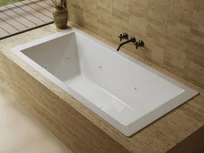 Modern white bathtub with sleek black faucet set in beige stone surround next to a window with potted plant.