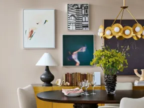 Modern dining room with round dark wood table, beige chairs, yellow sideboard, abstract art, and floral centerpiece.