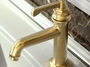 Brushed gold modern faucet mounted on white sink with dark marble backsplash in bathroom.