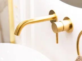 Modern bathroom faucet with gold finish mounted on white wall above white sink basin and beige towel.