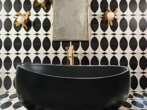 Modern black freestanding bathtub with gold faucet against black and white geometric tiled wall with decorative lights.