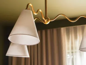 Modern ceiling light fixture with white cone-shaped shades and a wavy gold metal frame above beige curtains.