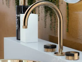 Modern gold faucet and matching basin with black and gold handles on white countertop with green plants background