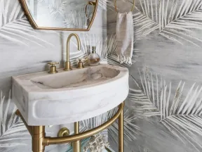 Modern bathroom with marble sink, gold fixtures, leaf-patterned wallpaper, and oval mirror under round light.