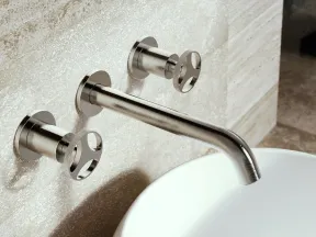 Modern stainless steel wall-mounted faucet with two handles above a round white sink on textured stone wall.