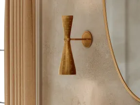 Modern brass wall sconce with perforated design mounted on textured beige wall beside large curved mirror.