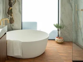 Modern bathroom with a round white bathtub, marble walls, wooden floor, glass shower, and decorative plant.