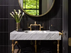 Modern bathroom with black tiled walls, round gold mirror, white marble sink, gold fixtures, and fresh flowers.