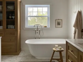 Cozy bathroom with freestanding white tub, wooden cabinets, stool with soap, window, and soft lighting.