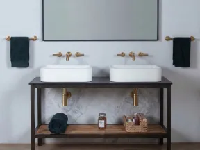 Modern double bathroom sinks with gold faucets, black towels, and wooden shelf with toiletries underneath.