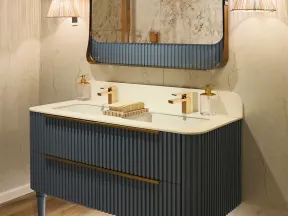 Modern bathroom vanity with ribbed dark blue cabinet, white countertop, dual gold faucets, and large mirror with lighting.