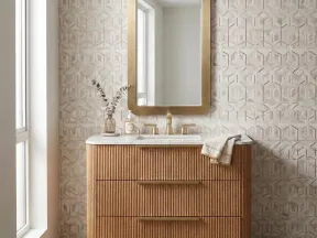 Modern bathroom vanity with wooden slatted drawers, brass faucet, large rounded rectangular mirror, and geometric tile wall.