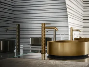 Modern bathroom sink with a gold faucet and round gold basin against a gray striped wall and mirror.
